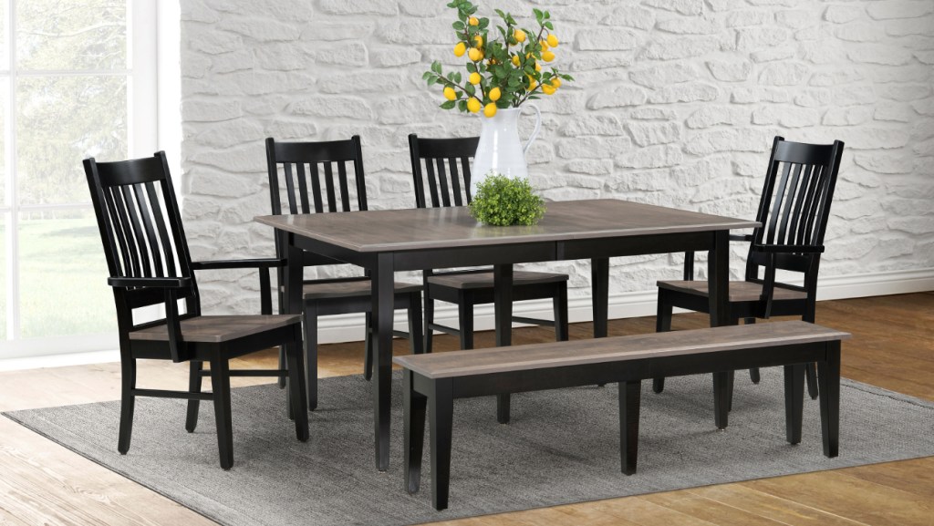 A modern farmhouse-style dining set featuring a rectangular wooden table with a dark finish, four matching slatted-back chairs, and a coordinating bench. The set is placed on a gray area rug against a white stone accent wall. A white pitcher filled with decorative lemon branches serves as the table’s centerpiece, adding a fresh, inviting touch. Large windows in the background allow natural light to brighten the space.