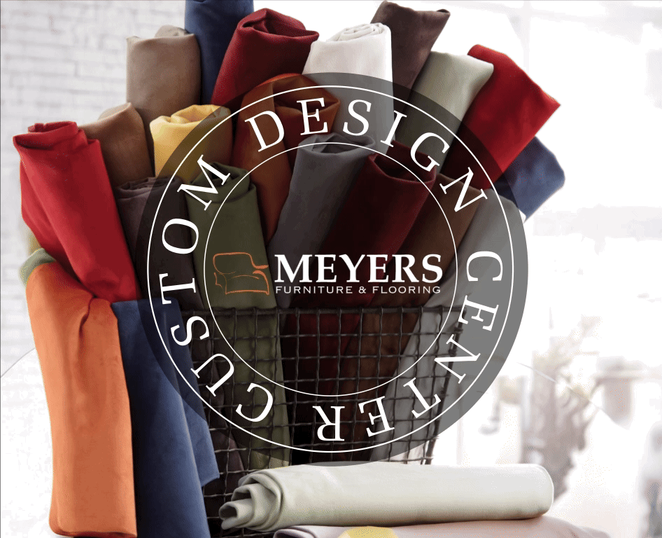 A basket filled with colorful fabric swatches, neatly rolled and arranged, showcasing a variety of hues and textures. Overlaid on the image is a circular logo with the words “CUSTOM DESIGN CENTER” surrounding the “MEYERS FURNITURE u0026 FLOORING” branding, which includes a stylized sofa icon. The background is softly blurred, emphasizing the vibrant fabric selection in the foreground.