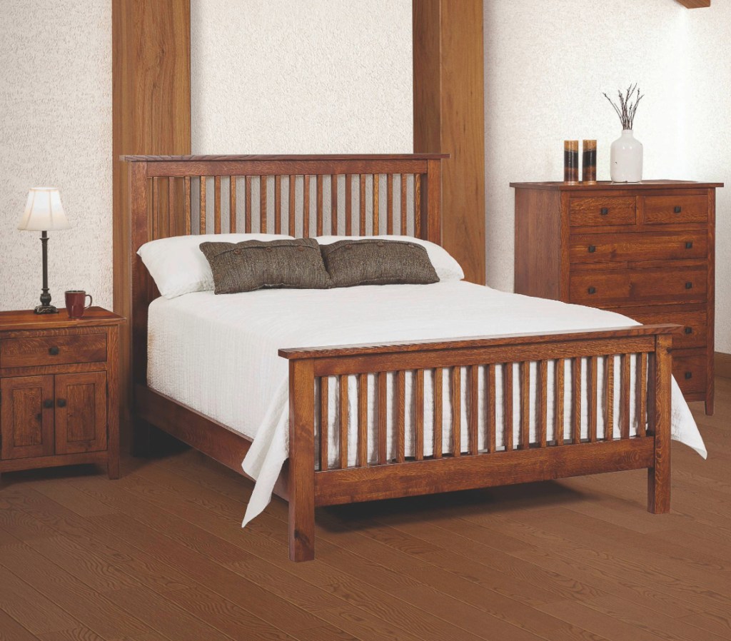 A classic Mission-style bedroom set crafted from rich, quarter-sawn white oak wood. The set features a slatted headboard and footboard bed, a matching nightstand with a lamp and mug, and a tall dresser adorned with decorative accents. The bed is dressed in crisp white linens with two accent pillows. The warm wood tones contrast beautifully with the neutral walls and textured backdrop, creating a timeless and inviting bedroom ambiance.