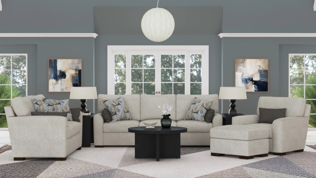 A modern living room featuring a neutral-toned sectional sofa with matching armchair and ottoman, adorned with patterned throw pillows. A round black coffee table sits in the center, flanked by two black side tables with table lamps. The walls are painted in a muted blue-gray tone, complemented by abstract artwork and large windows allowing natural light to filter in. A geometric area rug ties the space together.
