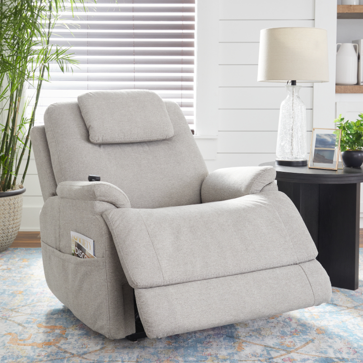 A modern light gray power recliner with an extended footrest is featured in a cozy living space. The chair includes a padded headrest, plush armrests, and a built-in side pocket for storing magazines. Positioned next to a black round side table, which holds a glass table lamp, a framed photo, and a small plant, the recliner sits on a patterned area rug. The background showcases white shiplap walls, a window with blinds, and a potted plant, adding to the inviting atmosphere.