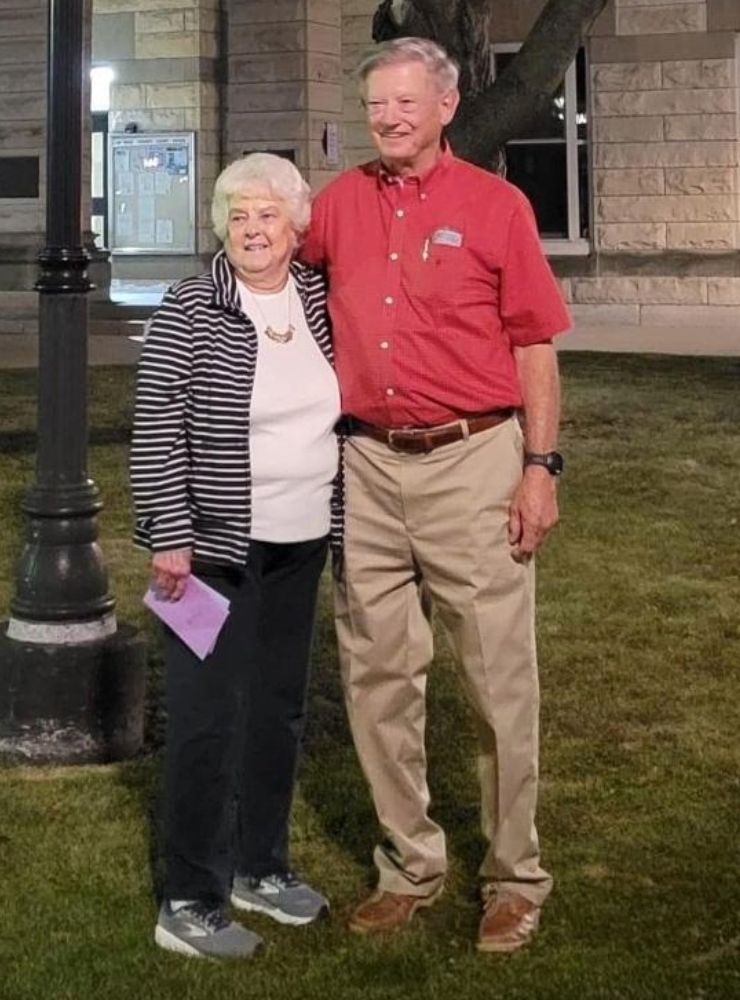 older couple posing together on a grassy area at night, with a historic stone building and lamppost in the background, both smiling and dressed casually.